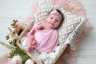 Newborn in pink wrap with a headband decorated with pale pink flowers lying in a basket on a crochet effect pillow, surround by flowers and foliage, by Jayne Poole Photography your newborn photographer near to me loacted in Great Torrington, near Fremington, Roundswell, Barnstaple.