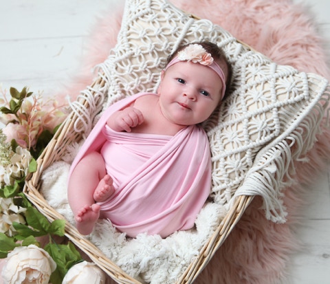 Baby in pink, with head band, with pale flower lying in a basket with cream crochet style pillow cover with flowers and foliage by Jayne Poole Photography based in North Devon